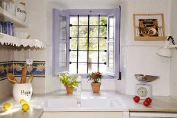 Mediterrane Küche mit weißem Spülbecken und mit Spitzen verziertem Wandboard, geöffnetes Fenster zeigt den Blick in den Garten