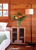 Kleiner Schrank mit Drahtgittertüren als Nachttisch neben Doppelbett; darauf ein prachtvoller Blumenstrauss mit orangen Blüten