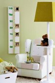 Kinderzimmer Feeling - Spielsachen auf Stehleuchte mit Ablage vor Polstersessel an grüner Wand mit Messlatte für Wachstumsschübe