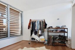 Clothes racks filled with stuff and shelves in a bedroom with large enclosed blind windows
