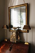 Antique clock flanked by lamps on top of bureau below gilt-framed mirror on wall