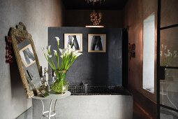 Graue Betontrennwand in Badezimmer mit schwarz gefliester Badewanne; im Vordergrund ein prachtvoller Calla-Strauss auf einem hellgrauen Beistelltisch