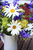 Sommerlicher Blumenstauß mit Margariten, blauen, roten und gelben Blütenpflanzen in hellgrauem Keramikkrug auf Gartenholztisch