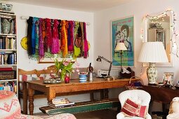 Zimmerecke mit Holzbank und Eichentisch, daneben ein volles Bücherregal und an der Wand eine Sammlung bunter Schals auf einer Vorhangstange trapiert; feminine Stimmung mit Lichterkette an Wandspiegel und Porzellantischlampe