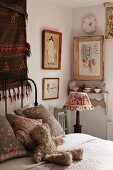 Corner of feminine bedroom with framed pictures and patterned china on wall bracket behind nostalgic bedside lamp on wooden chair; cuddly teddy bear waiting on cosy bed