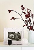 Romantischer weißer Bilderrahmen mit einer schwarz-weiß Fotographie von Hund mit Dame auf einem weiß lackierten Schrankelement mit schlichten weißen Milchglasblumenvasen daneben; ein rötlicher Zweig schenkt einen Farbakzent