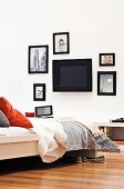 Black wall screen surrounded by pictures with black frames; cosy white bed with pillows and bedspread facing screen in foreground
