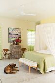 Bedroom in subdued shades with mosquito net above double bed with lime green throw; dog lying on cool stone floor