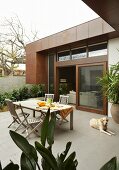 Erfrischungsgetränk auf Terrasse - Hund neben Tisch und Stühlen auf Fliesenboden vor Wohnhaus mit teilweise rostfarbener Metallverkleidung und offener Schiebetür