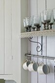Glasses on wall-mounted shelf with lace trim above cups hanging on rail in corner of white, wood-panelled kitchen