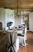 Rustikale Esstafel und Stühle mit Hussen aus grobem Leinen in gemütlichem Raum mit heller Holzdecke und großem Kerzenleuchter mit brennenden Kerzen