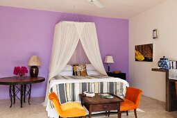 Double bed with net canopy and orange velvet chairs in lilac walled bedroom
