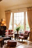 Wohnzimmer mit blumengemusterten Sesseln vor raumhohem Sprossenfenster; Im Vordergrund ein Polsterhocker aus braunem Leder