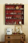 Various glasses and cups in wall-mounted shelving unit above vintage chest of drawers