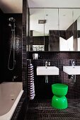 Modern bathroom with black mosaic tiles and green plastic stool in front of twin washbasins below mirrored cabinet