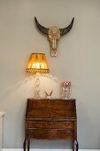 Table lamp on Baroque bureau against painted wall below animal skull