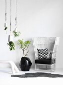 White wicker chair with black cushions, black floor vase against wall and decorative planters hanging from ceiling
