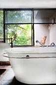 Freistehende Retro Badewanne mit Handtuchhalter vor Fenster mit halb geschlossener Jalousie und Blick in Garten