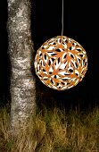 Spherical, perforated, orange pendant lamp hanging next to birch trunk against dark, night-time background like a lantern