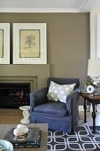 Comfortable, blue upholstered armchair and small, antique side table in front of modern open fireplace