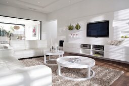 Weisses Wohnzimmer mit weisser Ledersofagarnitur und kreisförmige Couchtische auf flokatiartigem Teppich vor Sideboard unter Flachbild-Fernseher