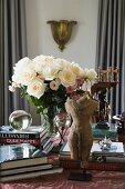 Miniature stone torso sculpture and books stacked around vase of white roses on table