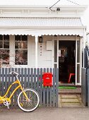 Entrance of white-painted cottage with porch; yellow bicycle leaning on grey garden fence with red letter box