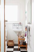 Einblick in kleines Badezimmer mit Holzregal und Waschbecken an weißer Wand, am Boden schwarz-weiß gefliestes Schachbrettmuster