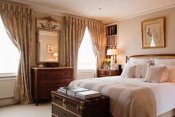 Antique chests of drawers, draped curtains and elegant trunk at foot of double bed in traditional bedroom
