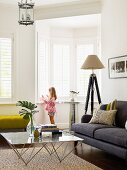 Glass coffee table and sofa with grey upholstery in corner of comfortable, elegant living room; little girl standing in bay window in background