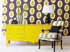 Flower arrangement and table lamp on yellow sideboard against wallpaper with brown and yellow pattern and behind comfortable upholstered armchair