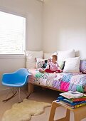 Kleines Kind auf Bett mit bunter Patchworkdecke, davor ein blauer Designer Schaukelstuhl
