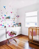 Klassiker Schaukelstuhl neben Kindergitterbett und weisses Sideboard an Wand mit Schmetterlingmotiven in sonnigem Kinderzimmer