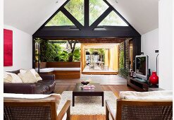 Blick vom Wohnraum mit Sitzbereich auf dreieckige Giebelverglasung, Terrasse mit Pergola und tropischen Garten