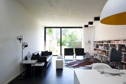Pale, modern interior with black leather couch, chrome standard lamp and long bookcase; sunny garden terrace in background