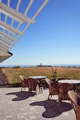 Rattanstühle und Tische auf sonnenbeschienener Terrasse unter Lamellen-Vordach vor weitläufiger Landschaft