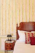Aussschnitt eines Bettes mit Kopfteil aus Holz und Intarsien neben antikem Nachtkästchen vor Wand mit Streifentapete