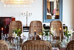 Glänzender Glastisch mit silbernen Kerzenhaltern und Liliendekoration; rundherum schalenförmige Rattanstühle