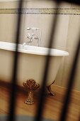 Blick durch unscharfe Gitterstäbe auf freistehende Badewanne mit Klauenfüssen und Wandfries aus Mosaikfliesen