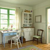 Halbhoch vertäfelter Wohnraum in Pastellfarben mit Vitrinenschrank und kleinem Schubladentisch vor grün lackierten Fensterrahmen
