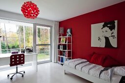 Teenager's bedroom with red wall, portrait of Audrey Hepburn, desk, red swivel chair and glass wall with view into garden