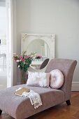 Cushions on dusky pink chaise longue, vase of flowers and oval mirror