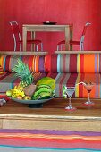 Outdoor furniture upholstered in bright stripes and bowls with tropical fruit on a wooden table; in the background a dining table in front of a bright, red wall