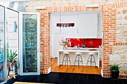 View from extension through wide, open doorway in brick wall into open-plan, white, designer kitchen with barstools at counter