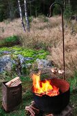 Rost mit Grillgut über loderndem Feuertopf auf einer Waldlichtung, daneben Blecheimer mit Brennholz