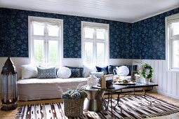 Gemütliches Wohnzimmer mit verschiedenen Beistelltischen vor langer Sitzbank am Fenster und weiße, halbhohe Holzverkleidung unter blauer gemusterter Tapete an Wand