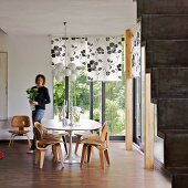 Seating area with 50s-style wooden chairs in corner of room with view of garden and woman carrying vase of flowers