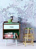 Small, fifties-style cabinet on legs next to stacked, yellow-painted metal stools against wall with crumbling plaster