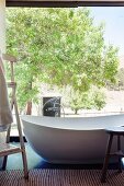 Free-standing designer bathtub in front of glass wall with view into sunny garden