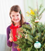 Mädchen steht mit Geschenk hinter Weihnachtsbaum